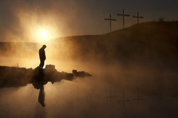 Becoming Gods Body of Love. Setting sun, silhouette of person reflected in a misty lake, three crosses visible on a distant hill.